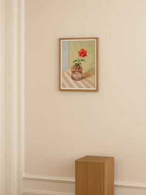 Framed artwork of a flower in a vase on a beige wall with a wooden pedestal.