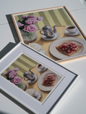 Two framed art prints featuring a table setting with a silver coffee pot, cup, whipped cream, crepe with strawberries, and flowers.