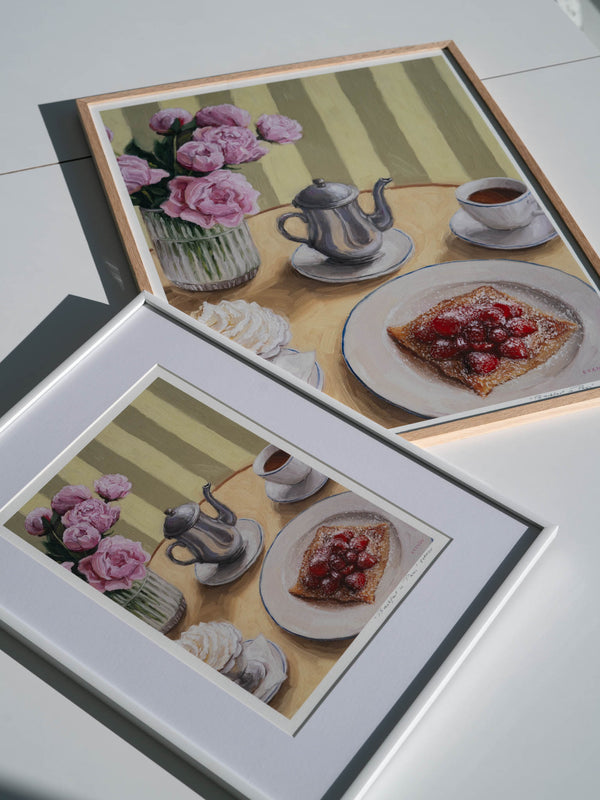 Two framed art prints featuring a table setting with a silver coffee pot, cup, whipped cream, crepe with strawberries, and flowers.